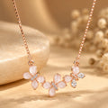 A rose gold necklace with a curved pendant featuring five pale pink flower petals and small clear crystal accents, displayed on a warm beige surface with soft blurred dried flowers in the background.